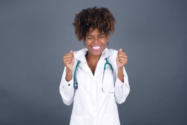  Afro-Amerikan doktor kadın doktor üniforması giyiyor başarısını ve zaferini kutluyor ve yumruklarını neşeyle sıkıyor. Hedeflerine ve hedeflerine ulaşmaktan mutlu olan şanslı kadın. Olumlu duygular, duygular.
