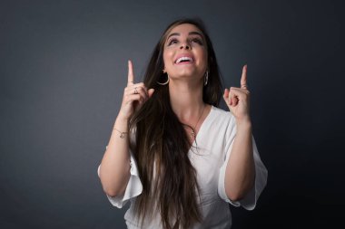 Olumlu ifadeli, kaygısız, çekici bir kadın, her iki işaret parmağıyla da işaret ediyor, günlük kıyafetler giymiş, pembe arka planda soyutlanmış, tedirgin bir gülümsemesi var. Şuraya bakın, lütfen. 