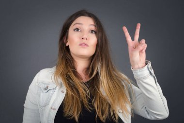 Beyaz kadın gri duvarın önünde duruyor. Bir numaralı parmaklarıyla işaret ediyor ve gülümsüyor. Kendinden emin ve mutlu..