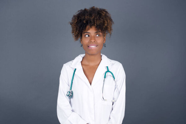 Dreamy  African American doctor female wearing medical uniform  with thoughtful expression, looks away keeps hands down bite her lip thinks about something pleasant poses against gray wall.