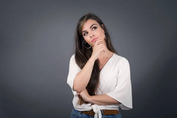 Portrait of thoughtful smiling girl keeps hand under chin, looks sideways, thinking or wondering about something with interest, dressed casually, poses against gray studio. Taking decisions concept.