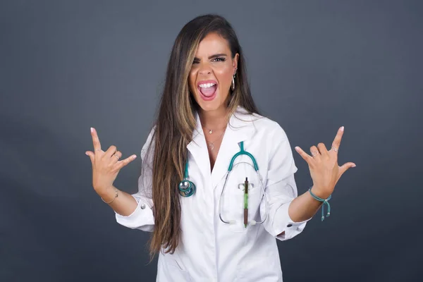 Born to rock this world. Joyful doctor woman in medical uniform screaming out loud and showing with raised arms horns or rock gesture, expressing excitement of being on concert of band.