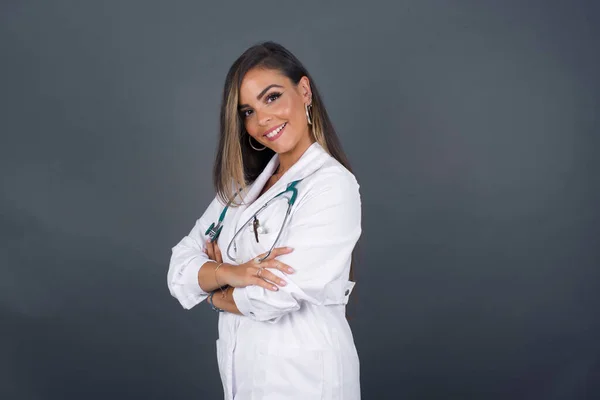 Image of cheerful pretty caucasian doctor lady standing against gray wall with arms crossed wearing medical uniform. Looking and smiling at the camera. Confident successful woman.