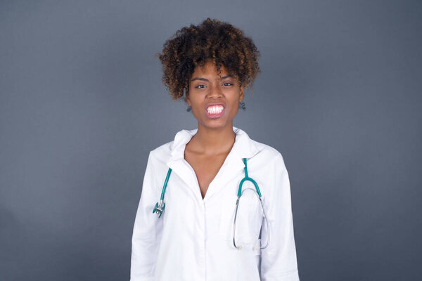 Mad crazy African American doctor woman clenches teeth angrily, being annoyed with coming noise, dressed in fashionable clothes, isolated over gray background. Negative feeling concept.