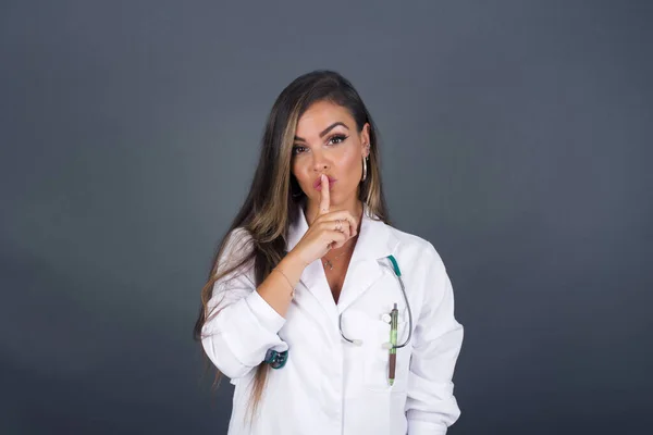 Don\'t tell my secret or not speak too loud, please! Emotional surprised young doctor woman wearing medical uniform makes hush gesture, asks be quiet.