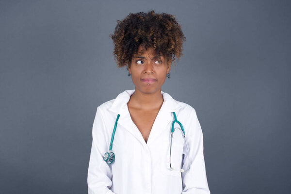 Photo of amazed puzzled  African American doctor female wearing medical uniform  curves lips and has worried look, sees something awful in front, isolated on white background, wearing medical uniform.