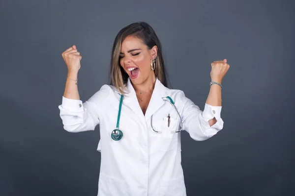 Caucasian brunette doctor woman wearing medical uniform and stethoscope, rejoicing her success and victory clenching her fists with joy happy to achieve her aim and goals. Positive emotions, feelings.