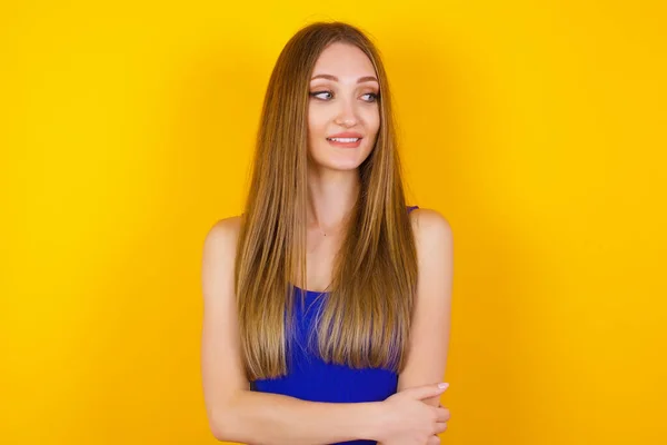 Dreamy young female with thoughtful expression, looks away keeps hands down bitting her lip thinks about something pleasant poses against gray wall.