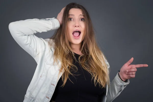 Portrait of young woman with shocked facial expression, showing something amazing on blank space, one hand on her head and pointing with forefinger. OMG concept.