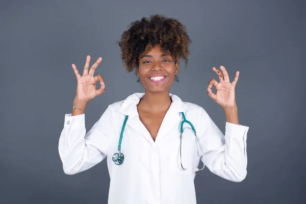 Glad attractive doctor woman shows ok sign with both hands as expresses approval, has cheerful expression being optimistic. Standing against gray wall.