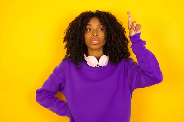 若い美しいアフリカ系アメリカ人女性は 紫色のニットセーターを着て黄色の背景を指し示しています — ストック写真