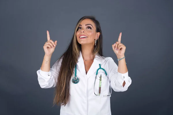 Horizontal shot of successful friendly looking young doctor woman with bright make up exclaiming excitedly, pointing both index fingers up, indicating something shocking on blank copy space wall.