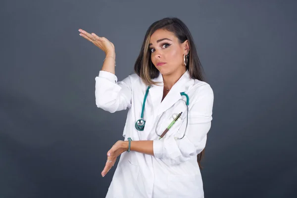 Beautiful European doctor girl pointing aside with both hands showing something strange and saying: I don\'t know what is this. Standing against gray background. Advertisement concept.