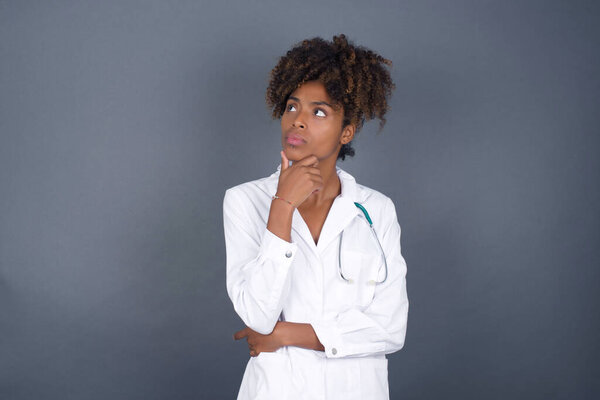 Portrait of thoughtful African American doctor woman wearing medical uniform keeps hand under chin, looks sideways, thinking or wondering about something with interest, dressed casually, poses against gray studio. Taking decisions concept.