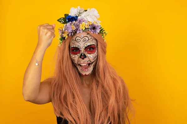 Fierce confident woman with halloween makeup holding fist in front of her as if is ready for fight or challenge, screaming and having aggressive expression on face. Isolated over yellow background.