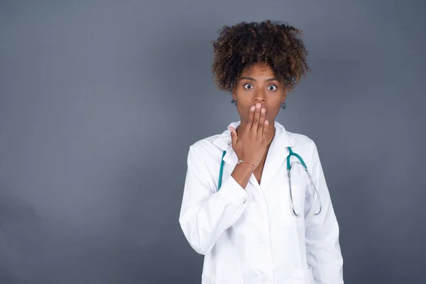 Oh! I think I said it! Close up portrait of doctor lady in white, covers open mouth by hand palm, looks at camera with big eyes isolated on bright mint background.