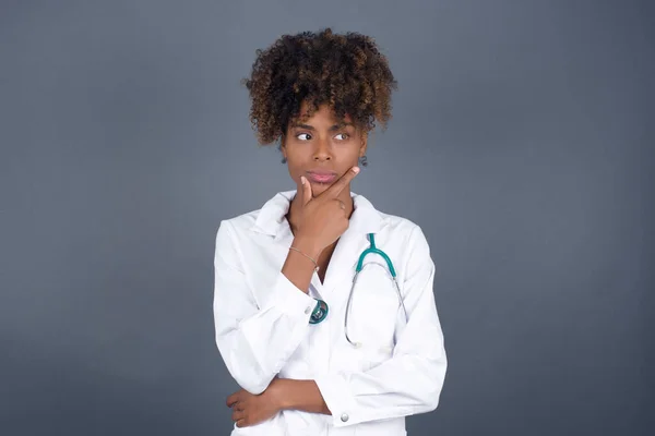 Isolated portrait of  African American doctor female wearing medical uniform  with hand under chin and looking sideways with doubtful and skeptical expression, worry and doubt. Standing indoors over gray background.