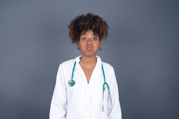Beautiful African American doctor woman puffing cheeks with funny face. Mouth inflated with air, crazy expression.