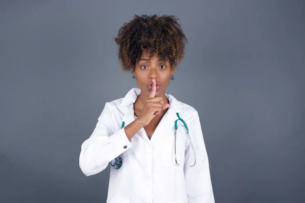 Don't tell my secret or not speak too loud, please! Emotional surprised young doctor woman wearing medical uniform makes hush gesture, asks be quiet.