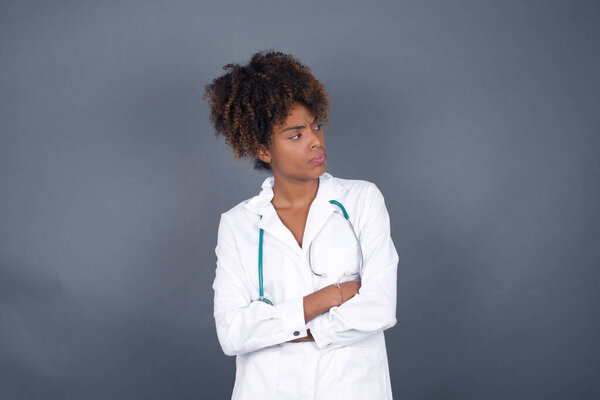 Image of upset African American doctor woman wearing medical uniform standing indoors with arms crossed. Looking with disappointed expression aside after listening to bad news. Confident girl.