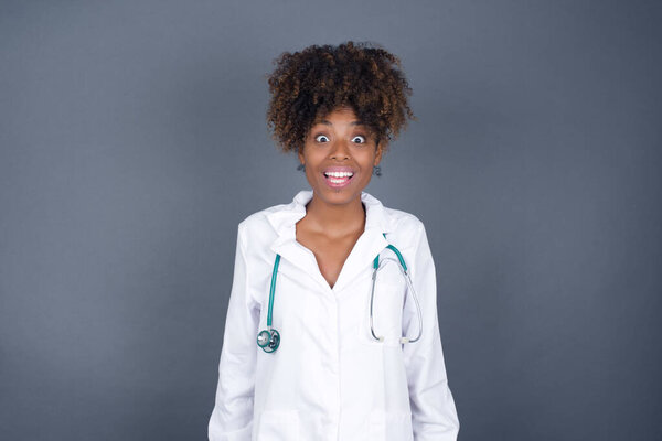 People and sudden reaction concept. Surprised African American doctor woman wearing medical uniform, shrugs shoulders, looking sideways, being happy and excited about new plans with friends. Isolated over gray wall.