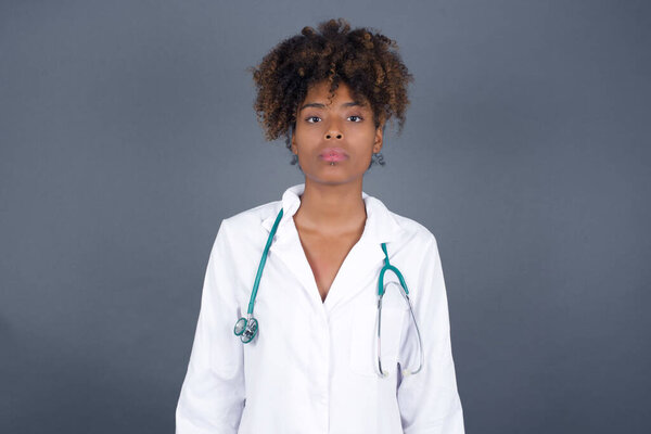 Waist up shot of joyful African American doctor woman wearing medical uniform looking to the camera, thinking about something. Both arms down, neutral facial expression wondering about something happening.