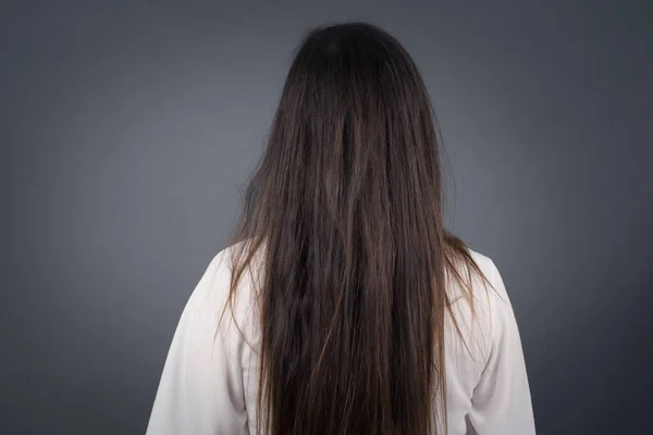 The back view of a girl with long straight and shiny hair standing against gray wall. Studio Shoot.