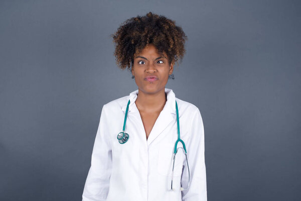 Funny comic African American doctor woman crosses eyes, with awkward expression has fun alone, puts lips, makes grimace, foolishes after all day.