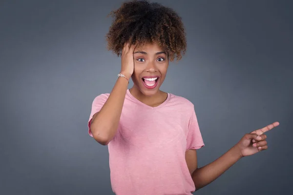 Portrait of young woman with shocked facial expression, showing something amazing on blank space, one hand on her head and pointing with forefinger. OMG concept.