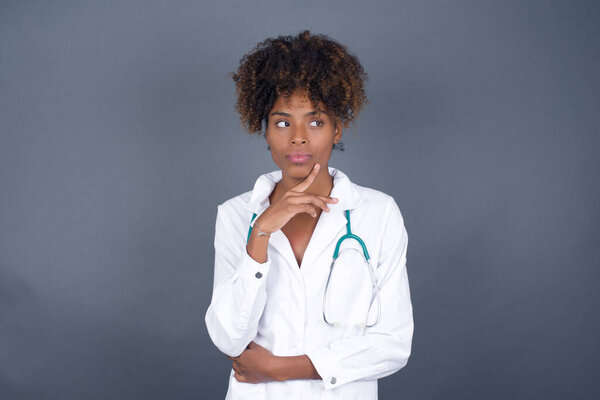 Dreamy African American doctor woman with pleasant expression, wearing medical uniform, looks sideways, keeps hand under chin, thinks about something pleasant, poses against gray background.
