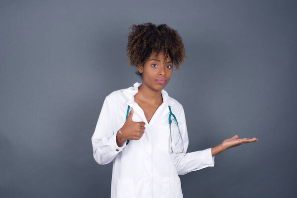 African American doctor woman wearing medical uniform showing thumb up and pointing with the other hand while standing indoors. Good Job. Wearing medical uniform.