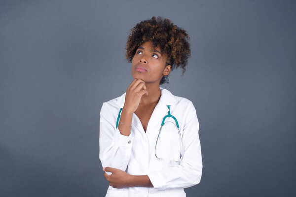 Portrait of thoughtful  African American doctor female wearing medical uniform  keeps hand under chin, looks sideways, thinking about something with interest, poses against gray studio. Taking decisions concept.