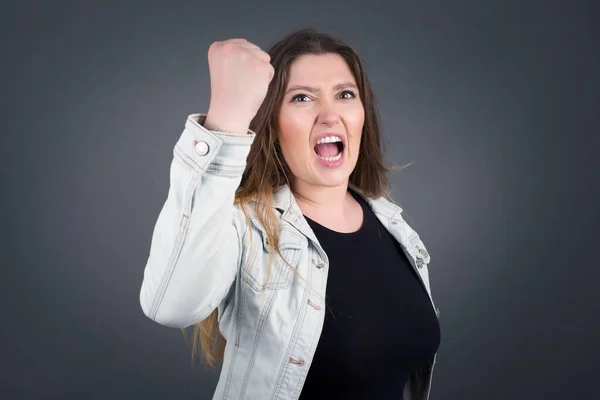 Fierce confident European dark-haired woman holding fist in front of her as if is ready for fight or challenge, screaming and having aggressive expression on face. Isolated over gray background.