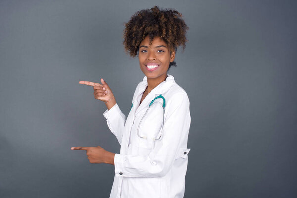 African American doctor woman wearing medical uniform, points aside with cheerful and surprised expression with mouth opened, shows something amazing standing indoors. Advertisement concept.