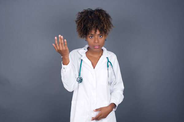 What the hell are you talking about, nonsense. Studio shot of frustrated African American doctor woman gesturing with raised palm, frowning, being displeased and confused with dumb question over gray wall.