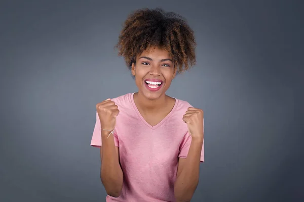 Waist up portrait of strong successful determined african american woman winner in casual t-shirt raising arms, clenching fists, exclaiming with joy and excitement. Victory, success and achievement concept.