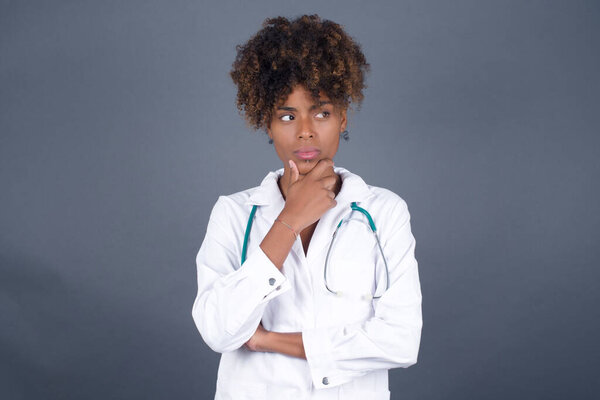 Portrait of thoughtful  African American doctor female wearing medical uniform  keeps hand under chin, looks directly at camera, listens to something with interest, dressed casually, poses against gray wall. Youth concept.