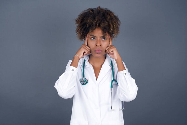 Dreamy African American doctor woman with thoughtful expression, looks away, keeps hand near face, thinks about something pleasant, poses against gray wall.