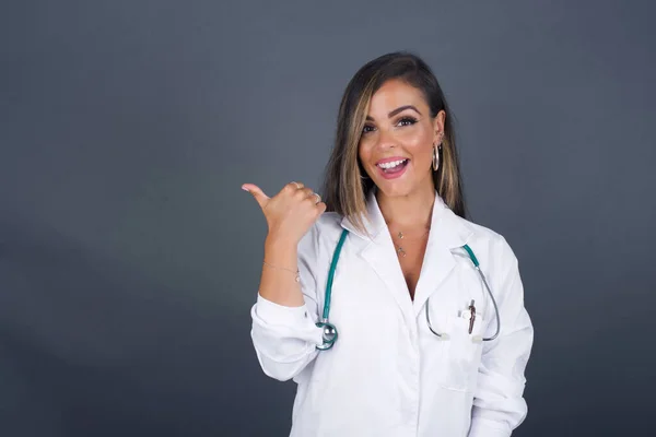 Omg concept. Stupefied caucasian doctor female with surprised expression, opens eyes and mouth widely, points aside with thumb, shows something strange on gray background. Advertisement concept.