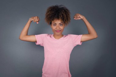 Genç ve güzel Afrikalı Amerikalı kadının belden yukarı çekimi kollarını kaldırarak kaslarının zaferden emin olduğunu, güçlü ve bağımsız göründüğünü, kameraya olumlu gülümsediğini, gri arka planda durduğunu gösterir. Spor konsepti.