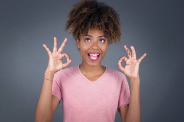 Çekici Afro-Amerikan kadınlarının tamam işareti göstermesine sevindim. Onayladıklarını belirterek, neşeli bir ifade takındılar. Güzel bir kadının fotoğrafı çekici bir görünüme sahip, iyimser olmak. Gri duvara yaslanarak.