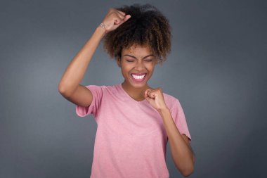 Genç ve güzel bir Afrikalı Amerikalı kadın zaferi kutluyor. Yumruklarıyla havaya yumruk atıyor. Gri bir stüdyo arka planında parlak dişli bir gülümsemeyle fotokopi çekiyor.