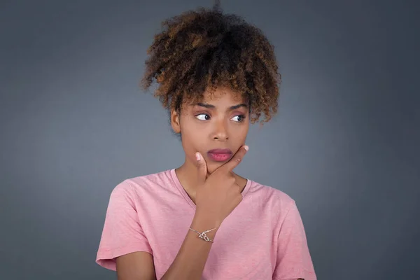 Isolated portrait of stylish young European woman with hand under chin and looking sideways with doubtful and skeptical expression, worry and doubt. Standing indoors over gray background.