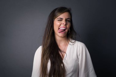 Gülümseyen ve dil gösteren mutlu ve komik yüzlü güzel beyaz kadın. Günlük giysiler giyip gri stüdyonun arka planında dikiliyorlar..