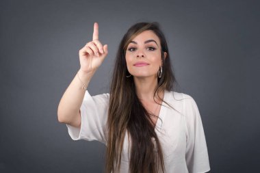 Beyaz kadın gri duvarın önünde duruyor. Bir numaralı parmaklarıyla işaret ediyor ve gülümsüyor. Kendinden emin ve mutlu..