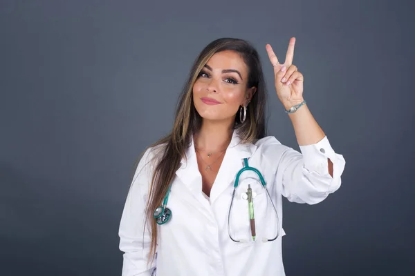 Joven Doctora Vistiendo Uniforme Médico Pie Contra Pared Gris Mostrando —  Fotos de Stock
