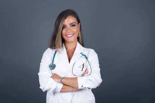 Kollarını kavuşturup gülümseyen mutlu yüzlü esmer doktor kız gri duvara karşı duran kameraya kendinden emin bir şekilde bakıyor. Pozitif kişi.