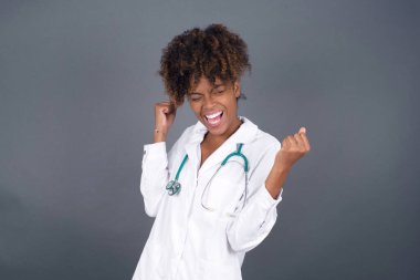 Afro-Amerikan kadın doktor, tıp üniforması giyip, başarısını ve zaferini kutluyor, yumruklarını neşeyle sıkıyor. Olumlu duygular, duygular.