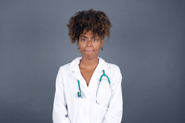 Pensive attractive african american woman doctor  in thoughts, raises eyebrows, curves lip. stands against gray wall with copy space for your text.
