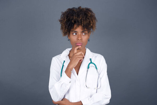Shot of contemplative thoughtful african american woman doctor  keeps hand under chin, looks thoughtfully upwards.